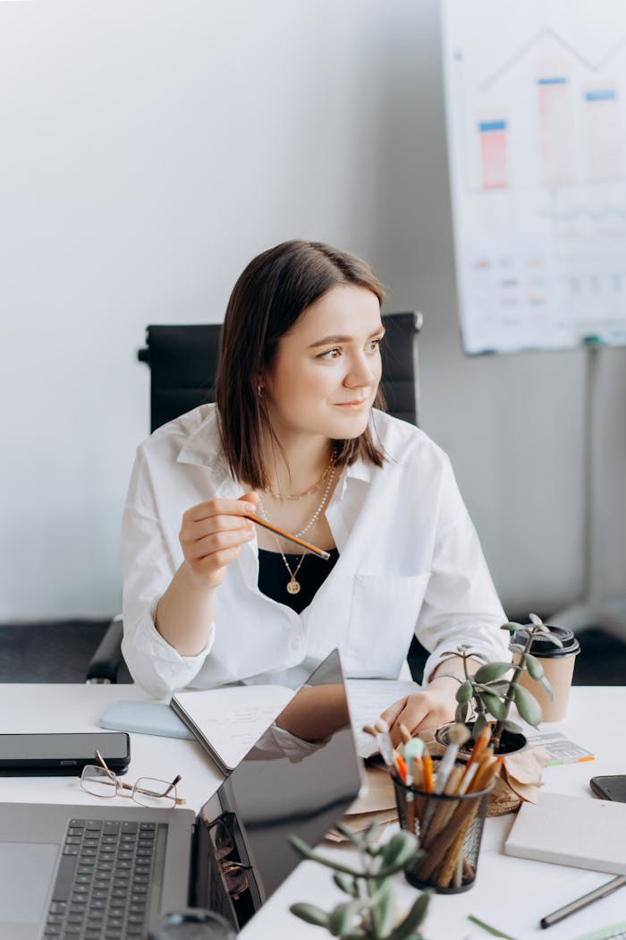 Businesswoman sits thoughtfully at desk with laptop and notes, projecting success and focus.